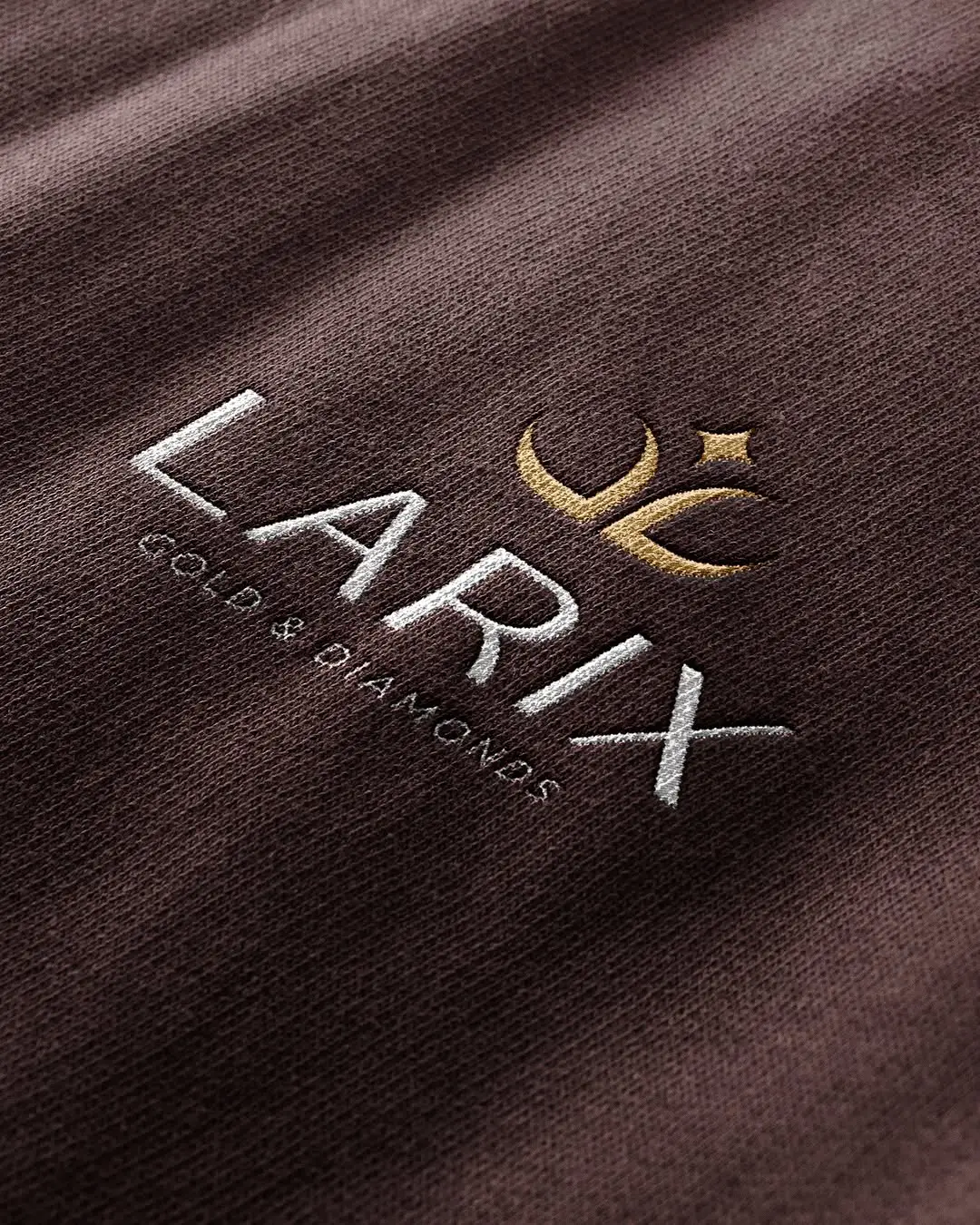 Larix logo embroidered in gold and white thread on plum fabric